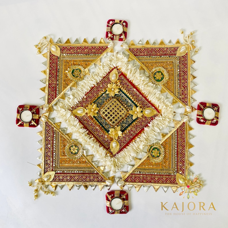 May include: A square-shaped decorative piece with intricate gold and red embroidery, surrounded by five small red candle holders. The center of the piece features a white, fluffy design with gold accents. The text "Kajara The House of Happiness" is visible at the bottom of the image.