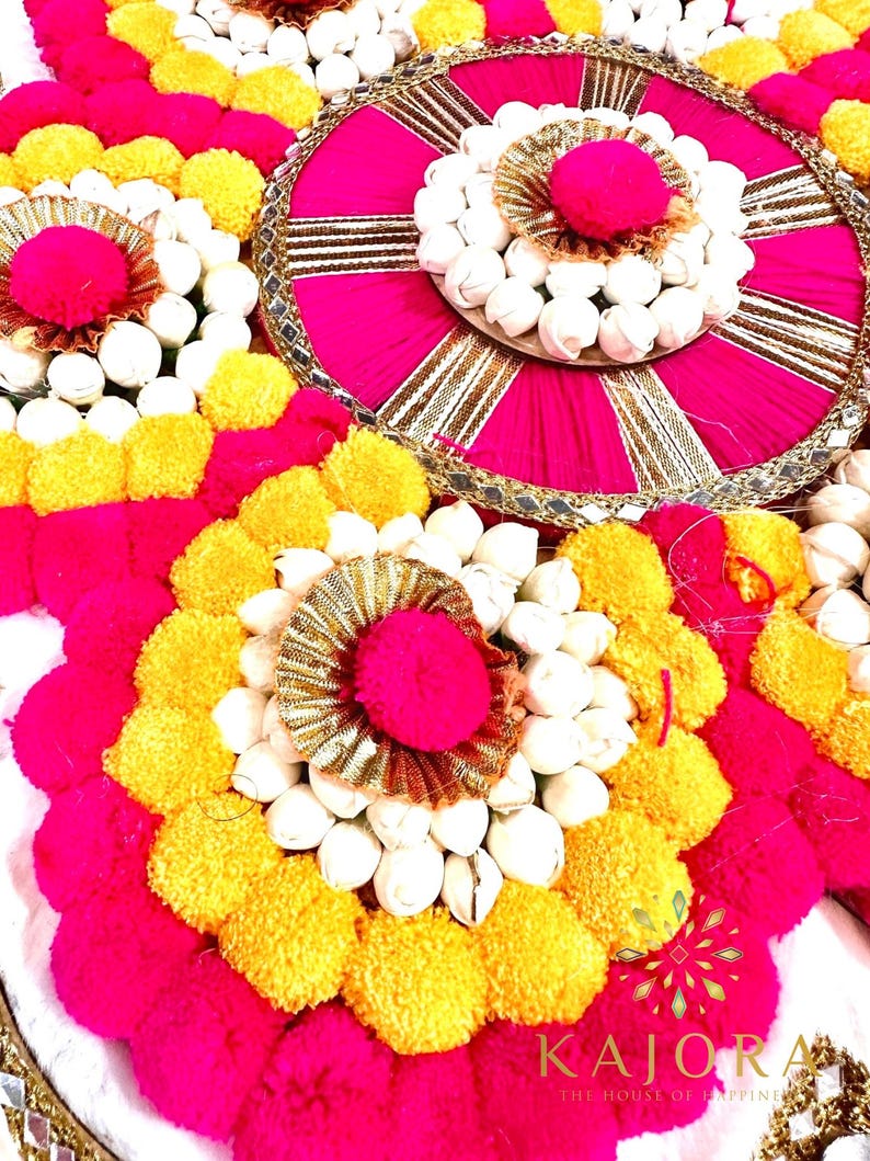 May include: A circular floral arrangement with pink and yellow pom-poms and white flowers. The center of the arrangement features a pink and gold circular design with a mirror-like surface.