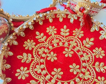 May include: A set of circular red velvet trays and a matching pouch featuring intricate gold embroidery, decorative gold trim, white pearl-like beads, and red tassels. The trays are adorned with floral patterns and a central red gemstone.