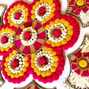 May include: A decorative floral arrangement with a circular design in the center. The arrangement is made of white, yellow, and pink pom-poms and is surrounded by smaller, similar arrangements. The text "KAJORA THE HOUSE OF HAPPINESS" is visible at the bottom of the image.