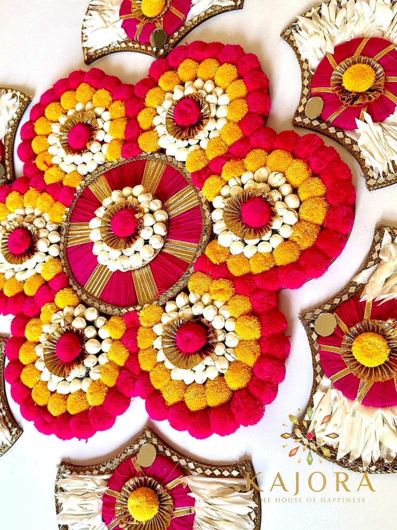 May include: A decorative floral arrangement with a circular design in the center. The arrangement is made of white, yellow, and pink pom-poms and is surrounded by smaller, similar arrangements. The text "KAJORA THE HOUSE OF HAPPINESS" is visible at the bottom of the image.