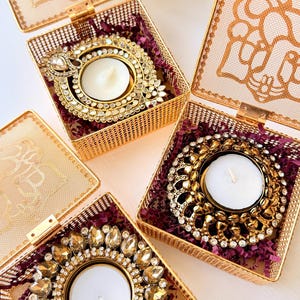 May include: Three square gold-toned metal mesh boxes with hinged lids, each containing a circular candle holder. The holders are decorated with clear and amber-colored crystals and rhinestones. Each box is lined with dark purple shredded paper.