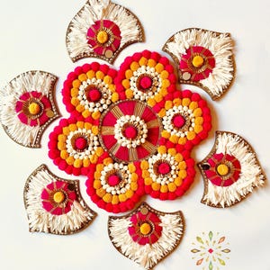 May include: A decorative wall hanging made of white, pink, and yellow pom-poms and fringe. The wall hanging is in the shape of a flower with a circular center.