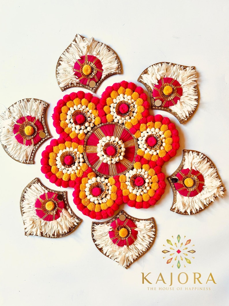 May include: A decorative wall hanging made of white, pink, and yellow pom-poms and fringe. The wall hanging is in the shape of a flower with a circular center.
