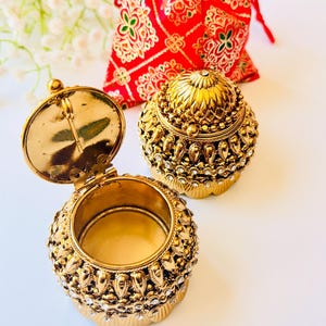 May include: Two small, round, gold-toned metal trinket boxes with intricate embossed patterns and embedded clear crystals. One box is open, revealing a smooth interior, while the other is closed with a domed lid. A red fabric pouch is behind them.