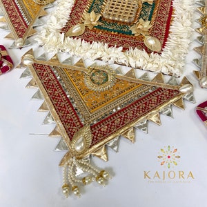 May include: A triangular decorative piece with a red, gold, and silver fabric design. The piece has a fringe of white pearls and is decorated with small mirrors and sequins.