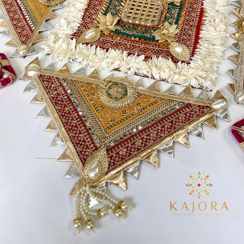 May include: A triangular decorative piece with a red, gold, and silver fabric design. The piece has a fringe of white pearls and is decorated with small mirrors and sequins.