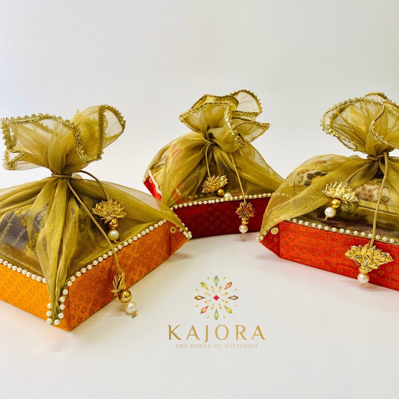Pu&ograve; includere: Tre piccole scatole regalo avvolte in tessuto arancione e rosso con bordi dorati. Ogni scatola &egrave; sormontata da un sacchetto di tessuto trasparente dorato e verde con un fiocco di perla.