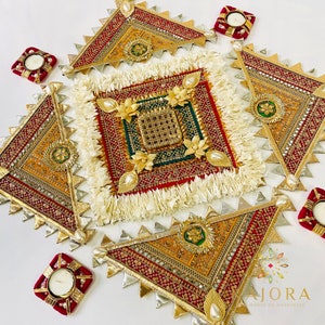 May include: A set of five decorative, triangular, gold and red fabric wall hangings with intricate embroidery and embellishments. The hangings are arranged in a square pattern with a smaller square in the center. The smaller square is decorated with white fringe and has a gold and green design. The hangings are surrounded by four small, red, square candle holders.