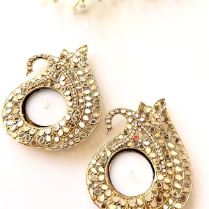 May include: Two gold-toned metal candle holders shaped like stylized birds, featuring intricate cut-out patterns and embedded reflective mirror pieces. Each holder has a circular center designed to accommodate a small round tea light candle.