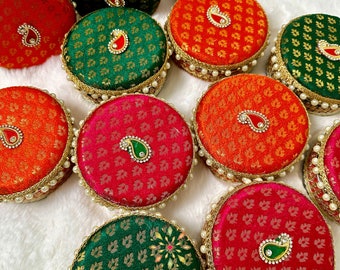 May include: Assortment of small round boxes with lids. The boxes are covered in fabric with a paisley pattern in shades of red, green, and orange. Each box has a small jeweled embellishment in the center.