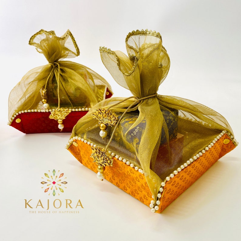 Pu&ograve; includere: Due scatole regalo quadrate avvolte in tessuto dorato e arancione con bordi dorati e dettagli perlati. Le scatole sono sormontate da sacchetti in tessuto dorato trasparente legati con un nastro dorato.