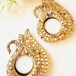 May include: Two gold-toned metal candle holders shaped like stylized peacocks, each featuring a circular center for a tea light candle. The surface of the holders is intricately decorated with numerous small, reflective mirror pieces and clear crystals.
