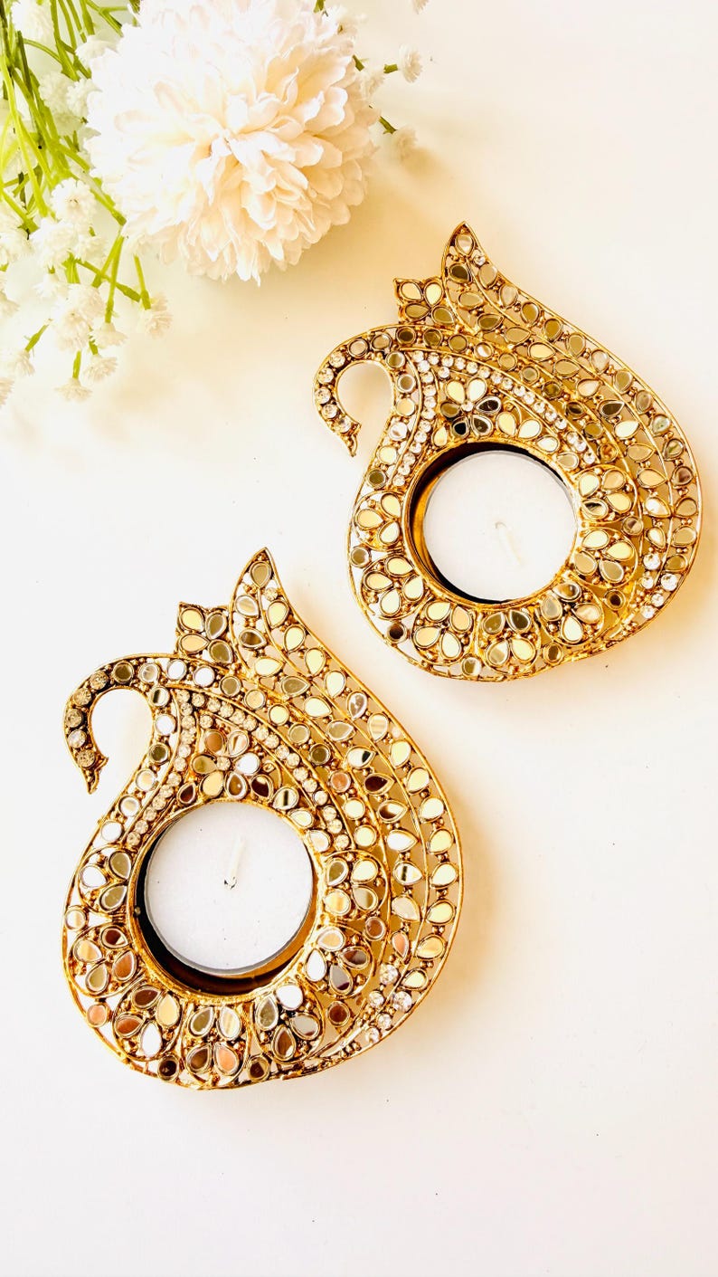 May include: Two gold-toned metal candle holders shaped like stylized peacocks, each featuring a circular center for a tea light candle. The surface of the holders is intricately decorated with numerous small, reflective mirror pieces and clear crystals.