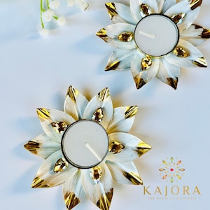 May include: Two flower-shaped candle holders with white petals featuring gold-painted tips and embedded amber-colored teardrop crystals. Each holder contains a round white tea light candle. The text KAJORA THE HOUSE OF HAPPINESS is visible.