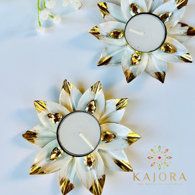 May include: Two flower-shaped candle holders with white petals featuring gold-painted tips and embedded amber-colored teardrop crystals. Each holder contains a round white tea light candle. The text KAJORA THE HOUSE OF HAPPINESS is visible.