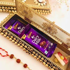May include: A rectangular gold-toned metal box with a mesh lid and decorative gold beads along the edges. Inside, two purple Cadbury Dairy Milk chocolate bars are placed alongside a small red and white decorative thread with a central gold accent.