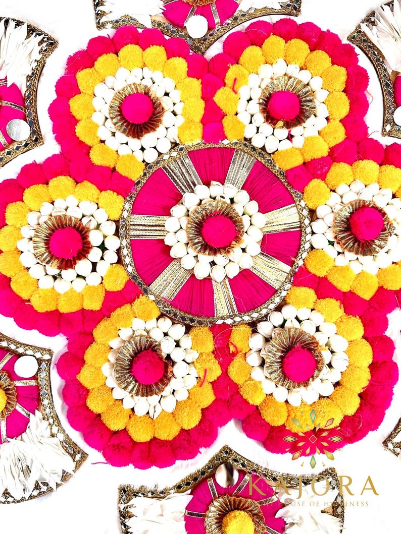 May include: A circular floral arrangement with pink, yellow, and white pom-poms. The arrangement is made up of several smaller circles, each with a different design. The center circle is pink with gold accents. The arrangement is surrounded by a white background.
