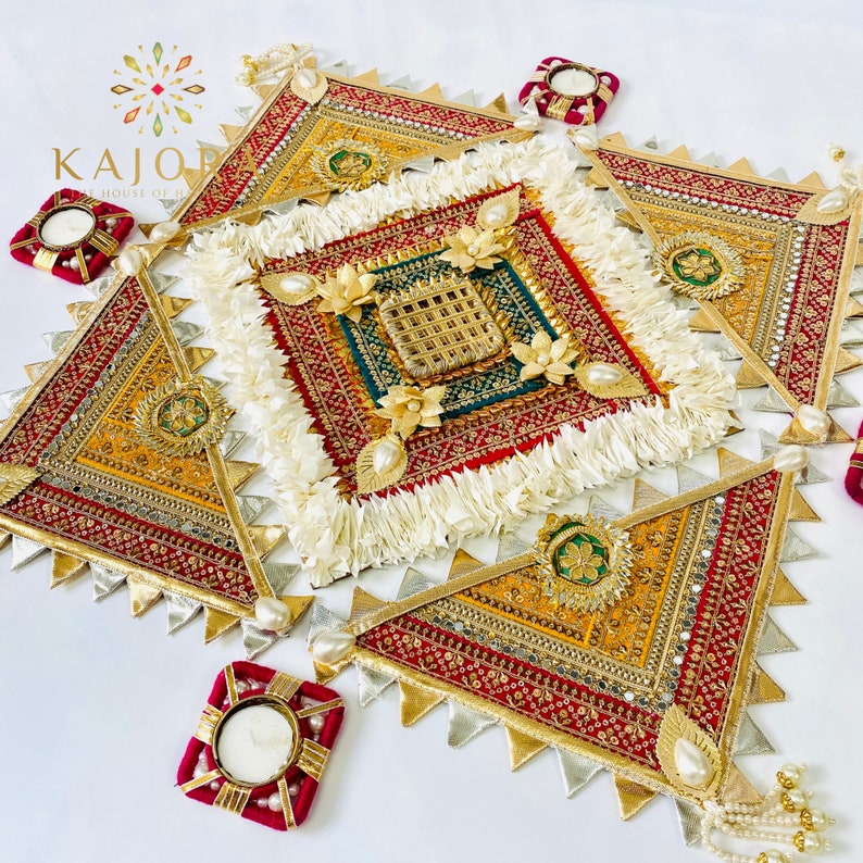 May include: A decorative wall hanging with a square design in the center, surrounded by four triangular pieces. The design features intricate embroidery with gold, red, and green accents. The center square has a white fringe border. The triangles have a silver and gold fringe border. The design is reminiscent of a traditional Indian mandala.