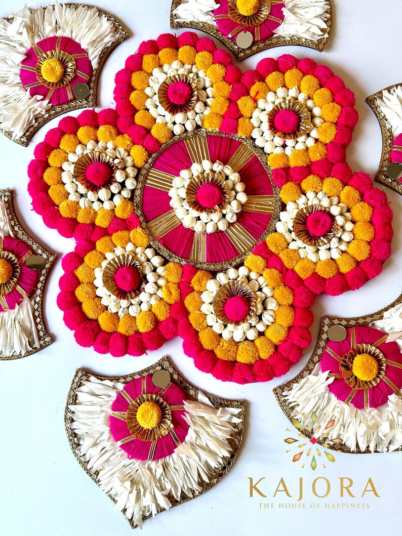 May include: A decorative wall hanging made of pink and yellow pom-poms, white fabric, and gold accents. The hanging is in the shape of a flower with a circular center and petals.