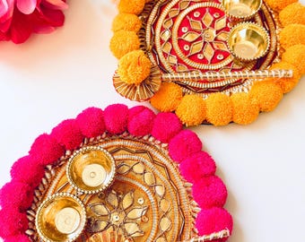 May include: Two circular decorative plates featuring vibrant pom-pom borders in orange and hot pink. Each plate displays intricate gold-toned metallic embroidery, a central floral motif, and two small gold-colored metal bowls with a matching stick.