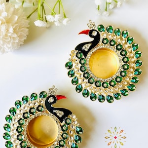 May include: Two circular candle holders shaped like peacocks, featuring black enamel heads with red beaks, gold-toned centers, and decorative borders of green and teal teardrop-shaped stones accented with clear crystals. The text KAJORA is visible.