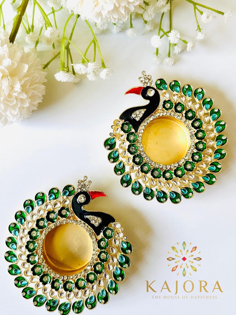 May include: Two circular candle holders shaped like peacocks, featuring black enamel heads with red beaks, gold-toned centers, and decorative borders of green and teal teardrop-shaped stones accented with clear crystals. The text KAJORA is visible.
