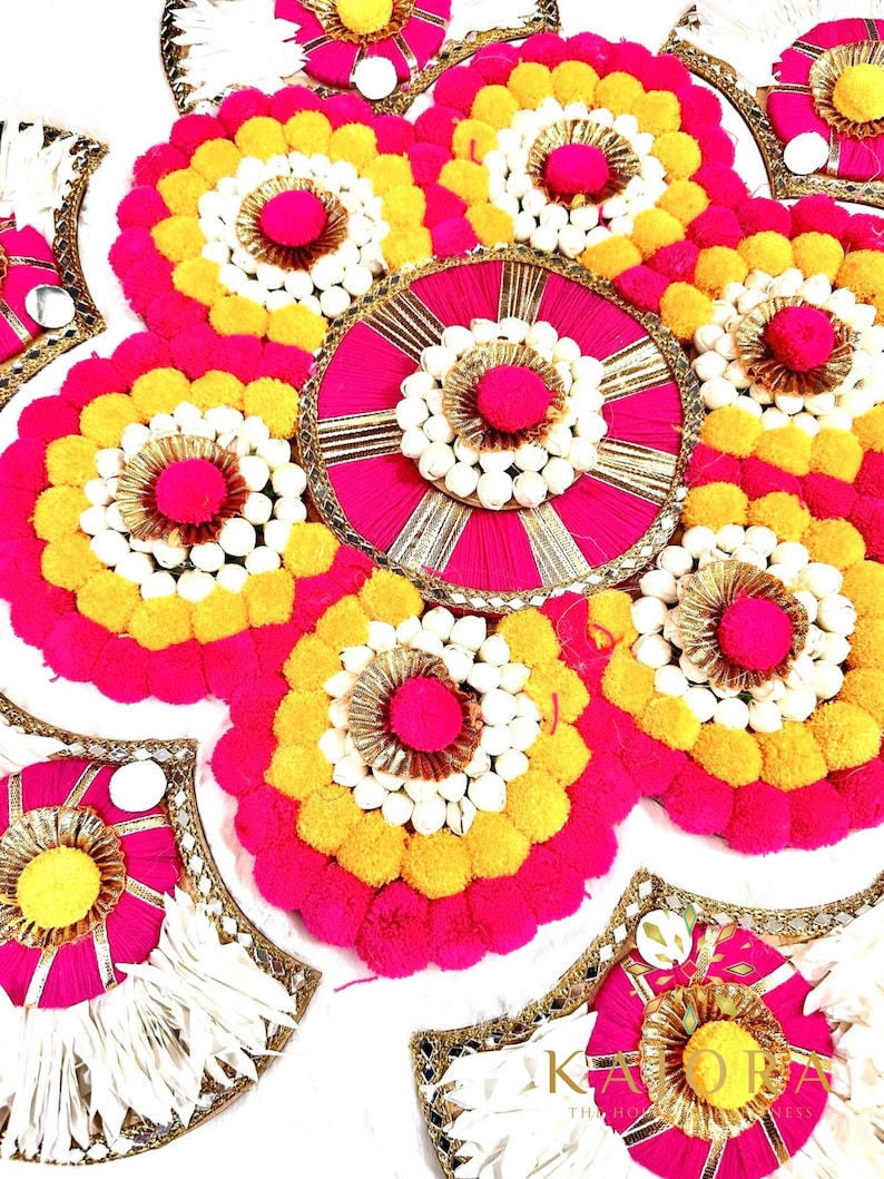 May include: A set of decorative round and fan-shaped floral arrangements. The arrangements are made with pink, yellow, and white pom-poms and are accented with gold trim. The arrangements are on a white background.