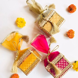 May include: Four decorative gift boxes with gold, yellow, and pink fabrics. Each box is square-shaped, adorned with gold trim, tassels, and tied with a ribbon. Scattered around are yellow and orange flowers, creating a festive arrangement.