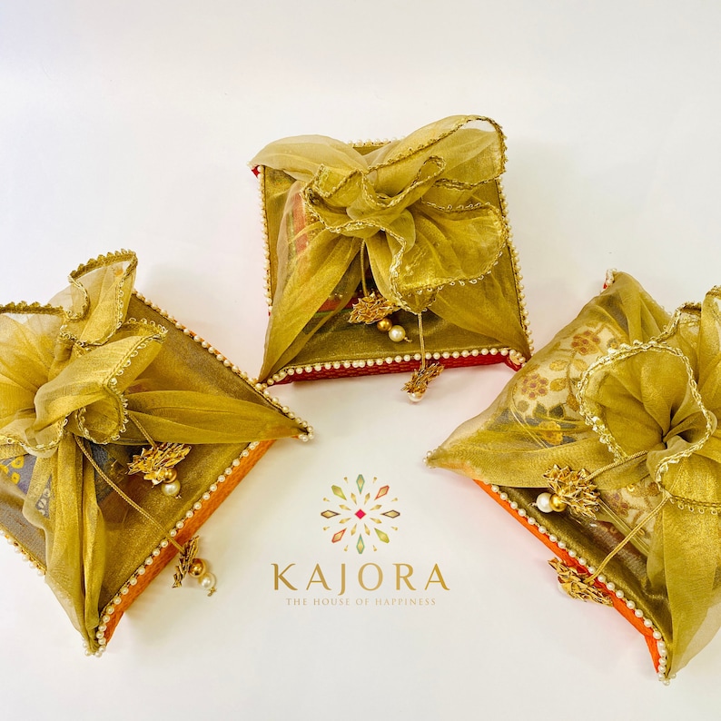 Pu&ograve; includere: Tre scatole regalo quadrate avvolte in tulle dorato con bordi dorati e accenti di perle bianche. Le scatole sono su una superficie bianca. Le scatole sono decorate con un bordo rosso e arancione. Il testo "Kajora The House of Happiness" &egrave; visibile sotto le scatole.