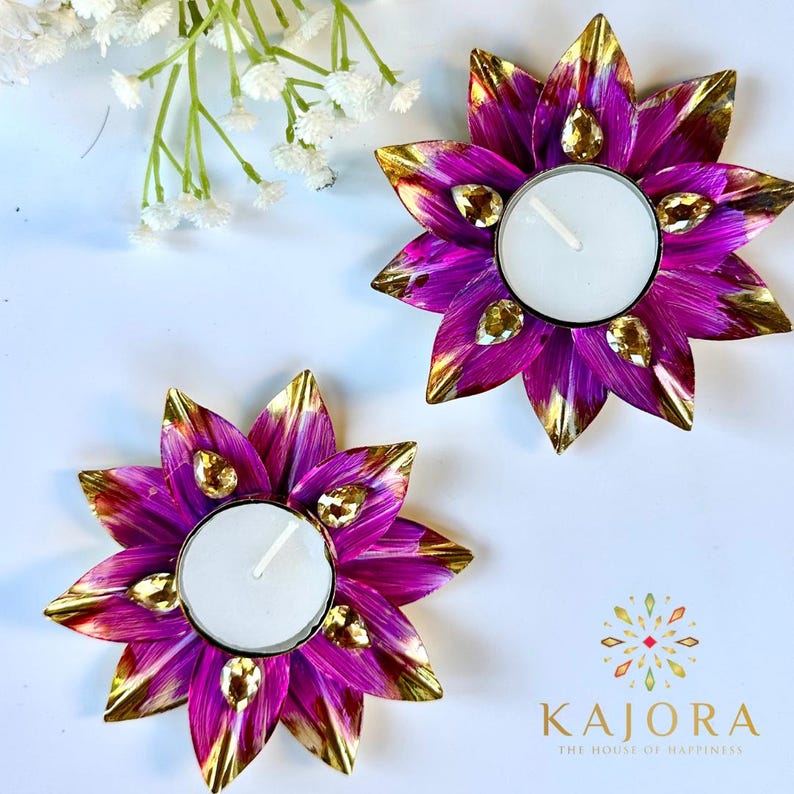 May include: Two flower-shaped candle holders with vibrant purple petals featuring gold-toned tips and embedded clear teardrop-shaped crystals. Each holder contains a round white tea light candle. The logo KAJORA THE HOUSE OF HAPPINESS is visible.
