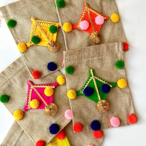 May include: A collection of burlap drawstring bags decorated with colorful, kite-shaped embellishments. Each kite is a different color, including yellow, orange, pink, and green, and adorned with matching pom-poms. The bags are ideal for gifts or small items.