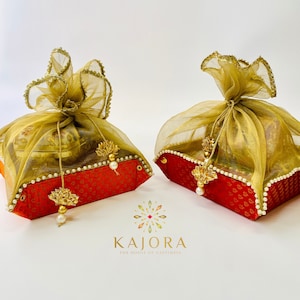 Pu&ograve; includere: Due scatole regalo rosse con bordi dorati e dettagli di perle sono avvolte in tessuto trasparente dorato e verde. Le scatole sono legate con un fiocco e hanno un nappa dorata appesa a ciascuna. Le scatole sono su una superficie bianca. Il testo "Kajora The House of Happiness" &egrave; stampato sotto le scatole.