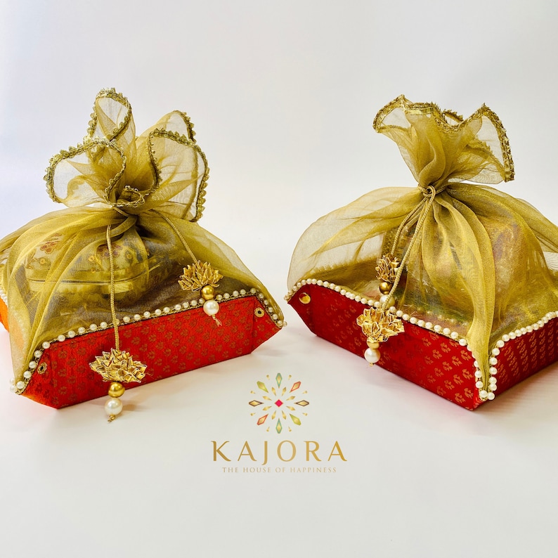 Pu&ograve; includere: Due scatole regalo rosse con bordi dorati e dettagli di perle sono avvolte in tessuto trasparente dorato e verde. Le scatole sono legate con un fiocco e hanno un nappa dorata appesa a ciascuna. Le scatole sono su una superficie bianca. Il testo "Kajora The House of Happiness" &egrave; stampato sotto le scatole.