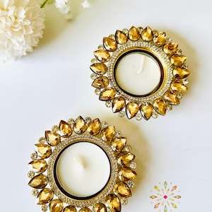 May include: Two circular candle holders featuring a central white tea light candle surrounded by a ring of amber-colored, teardrop-shaped crystals and a border of small clear rhinestones. The text KAJORA THE HOUSE OF HAPPINESS is visible.