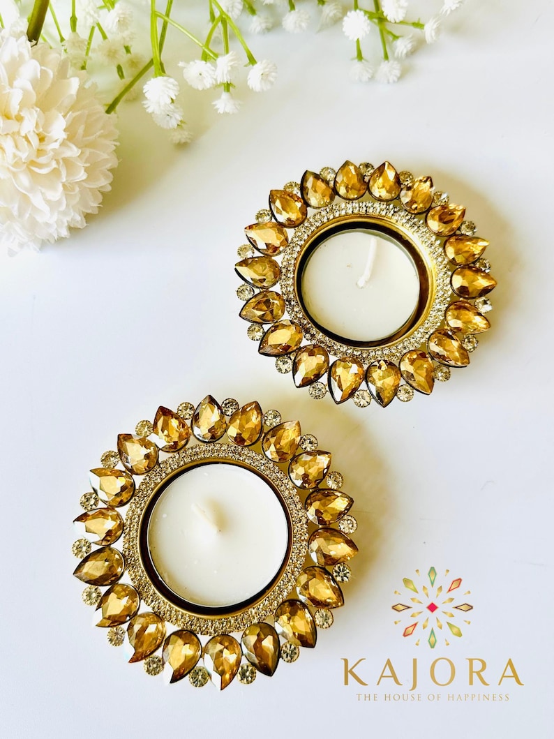 May include: Two circular candle holders featuring a central white tea light candle surrounded by a ring of amber-colored, teardrop-shaped crystals and a border of small clear rhinestones. The text KAJORA THE HOUSE OF HAPPINESS is visible.