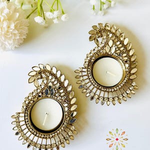 May include: Two gold-toned metal tea light candle holders shaped like paisley motifs, embellished with small clear crystals and reflective mirror pieces. Each holder features a central circular cup for a candle. The text KAJORA THE HOUSE OF HAPPINESS is printed in the bottom right corner.