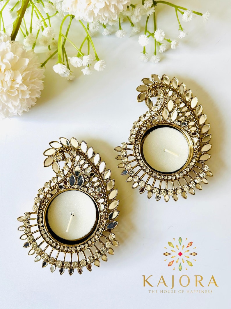 May include: Two gold-toned metal tea light candle holders shaped like paisley motifs, embellished with small clear crystals and reflective mirror pieces. Each holder features a central circular cup for a candle. The text KAJORA THE HOUSE OF HAPPINESS is printed in the bottom right corner.