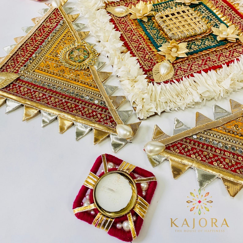 May include: Three decorative, triangular, fabric wall hangings with gold, red, and silver embellishments. The hangings are arranged in a triangular shape. A small, square, red fabric candle holder with gold trim and white beads is in the center of the image. The text "Kajara The House of Happiness" is in the lower right corner of the image.