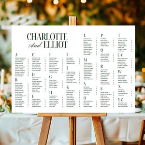 May include: A white seating plan on a wooden easel, with the names "Charlotte" and "Elliot" at the top. The chart is organised alphabetically with guest names listed under each letter. The background is blurred with soft lighting.