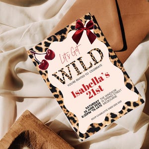 May include: A party invitation with a leopard print border and the words "Let's Get WILD" in bold letters. The invitation is for Isabella's 21st birthday, with details including the date, time, and location. A red bow and cherries add a decorative touch.