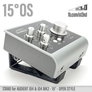 May include: A silver-coloured audio interface with a black stand. The stand is angled at 15 degrees. The interface has a volume knob, mute button, and several other knobs. The text "audient" is visible on the interface. The text "15°0S" is visible in the top left corner of the image. The text "STAND for AUDIENT ID4 & ID4 MK2 - 15° - OPEN STYLE" is visible at the bottom of the image.