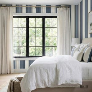May include: A bedroom interior featuring a large window, white curtains, and a bed with white linen bedding. The walls are adorned with vertical blue and white striped wallpaper. A woven basket is near the bed.