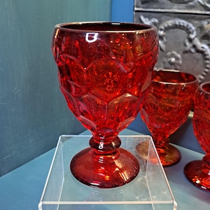 May include: Three red glass goblets with a textured, faceted design. The goblets are on a clear acrylic display stand.