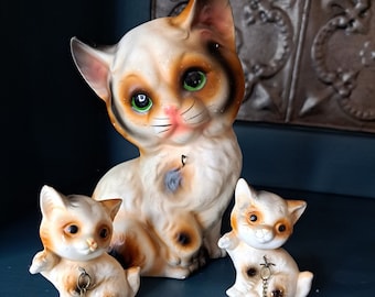 May include: Three ceramic cat figurines, a larger white cat with brown and orange markings and two smaller white cats with brown markings. The cats are connected by gold chains.