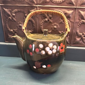 May include: A vintage green ceramic teapot with a woven handle and lid. The teapot is decorated with red and white floral designs. The teapot is sitting on a blue surface.