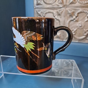 May include: Black ceramic mug with a gold and white crane design. The mug has a brown rim and handle.