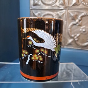 May include: Black ceramic cup with a gold and white crane design. The crane is flying over a branch with orange and green flowers.