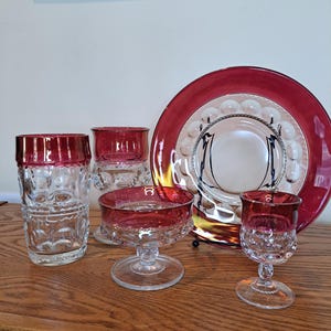 May include: Set of five vintage glass pieces with a ruby red rim and clear glass body. The set includes a large plate, a tall glass, a shorter glass, and two stemmed glasses.