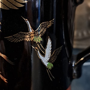 May include: Black ceramic mug with a gold and white crane design. The crane is flying with its wings spread. The mug has a brown rim.